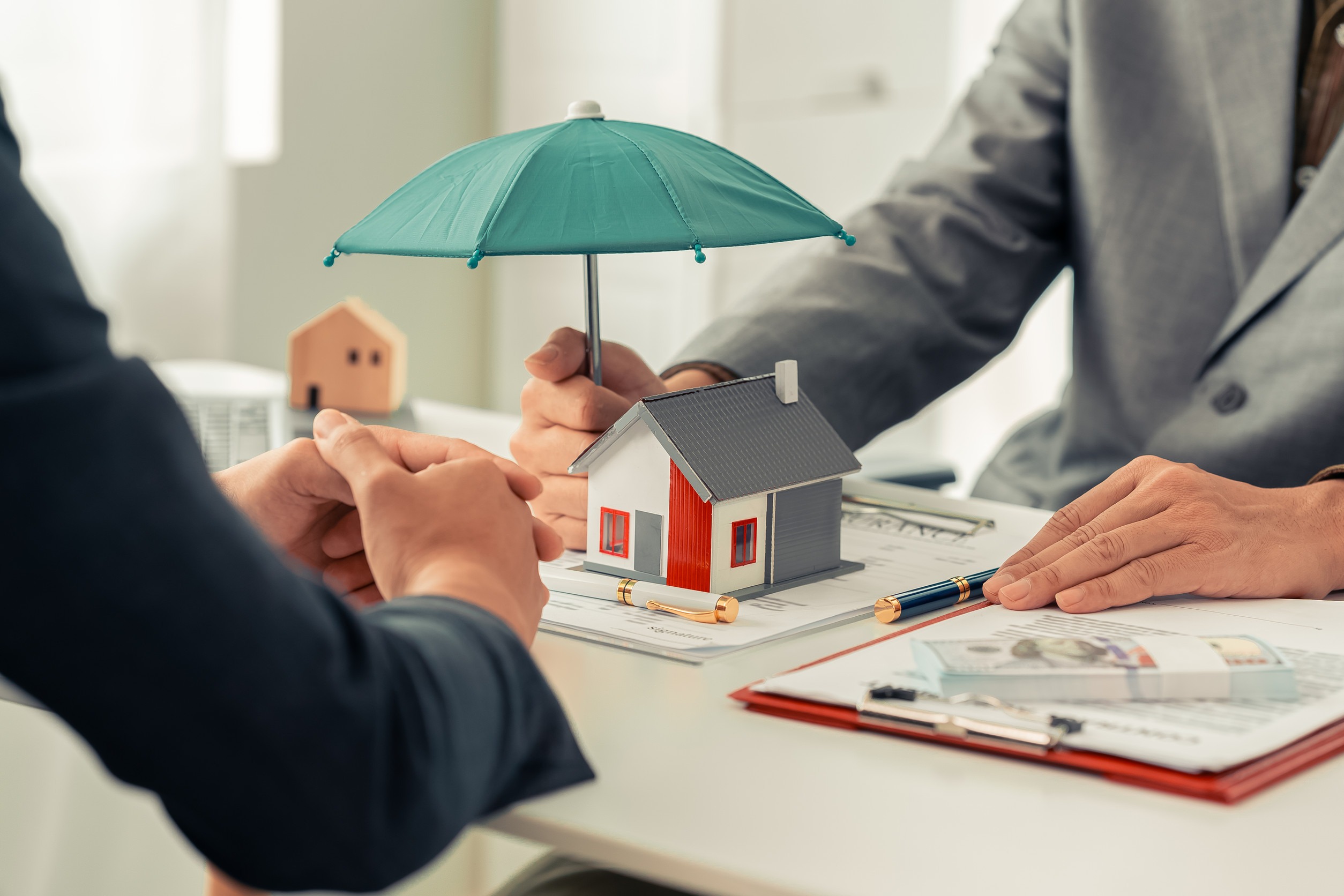 Two people sit at a desk discussing homeowners insurance. One holds a model house while the other places a green umbrella over it, symbolizing protection and insurance coverage. Documents and clipboards are visible on the table.