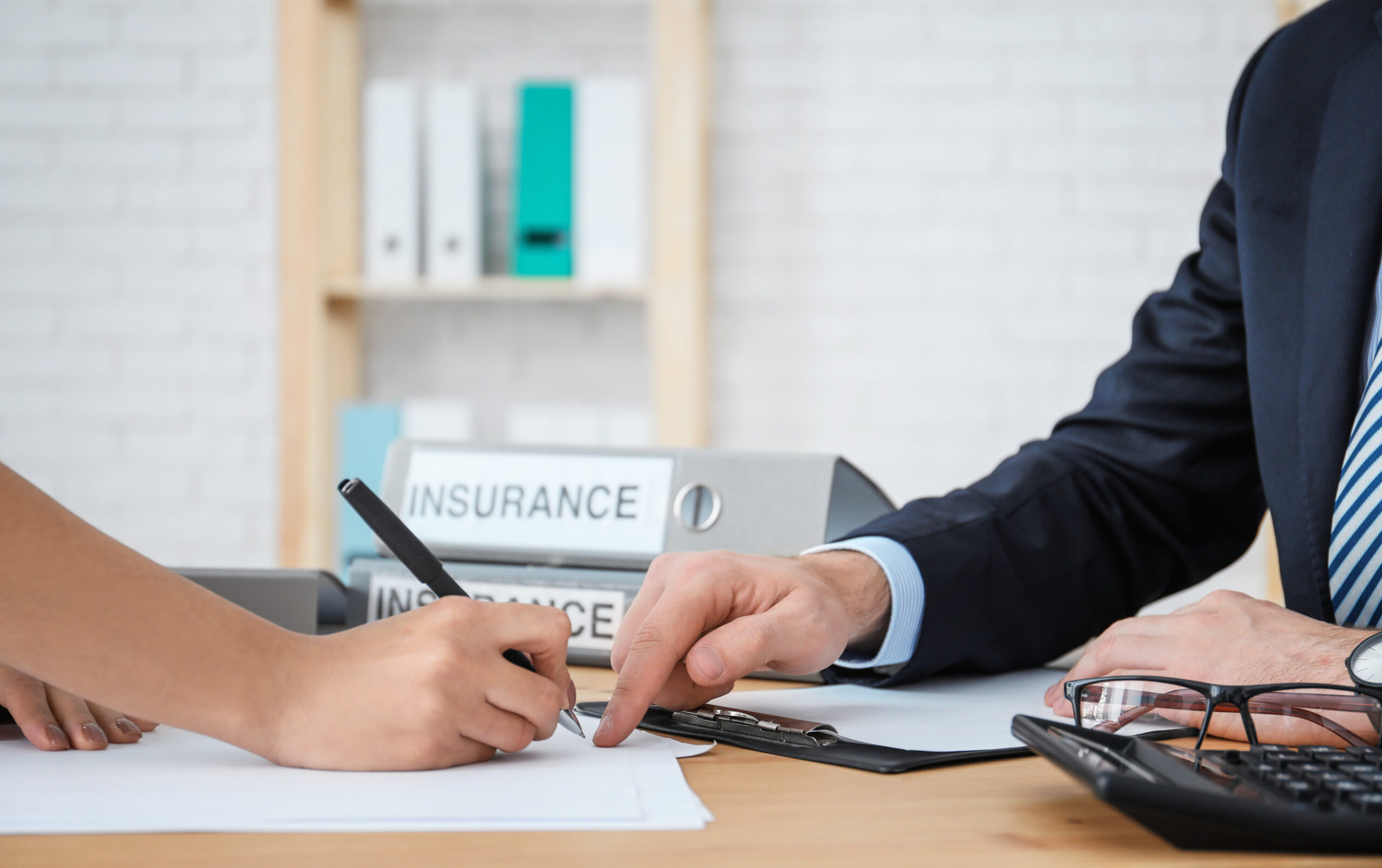 A person is signing a document on a desk while an independent insurance broker in a suit points to the paper. There are folders labeled 