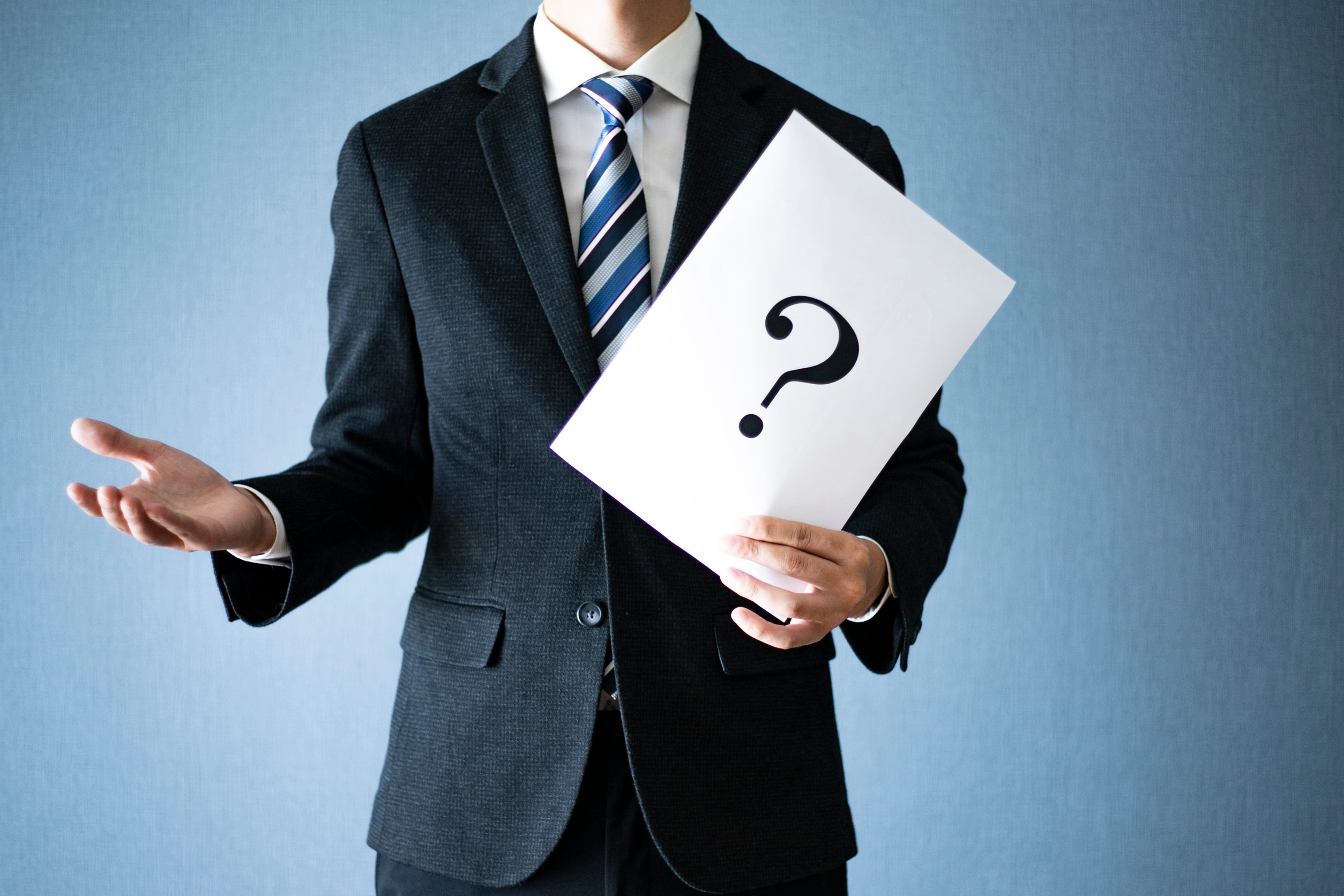 A person in a suit and striped tie holds a white paper with a large black question mark on it, hinting at uncertainty about insurance deductibles, standing against a plain blue background. The person's face is not visible.