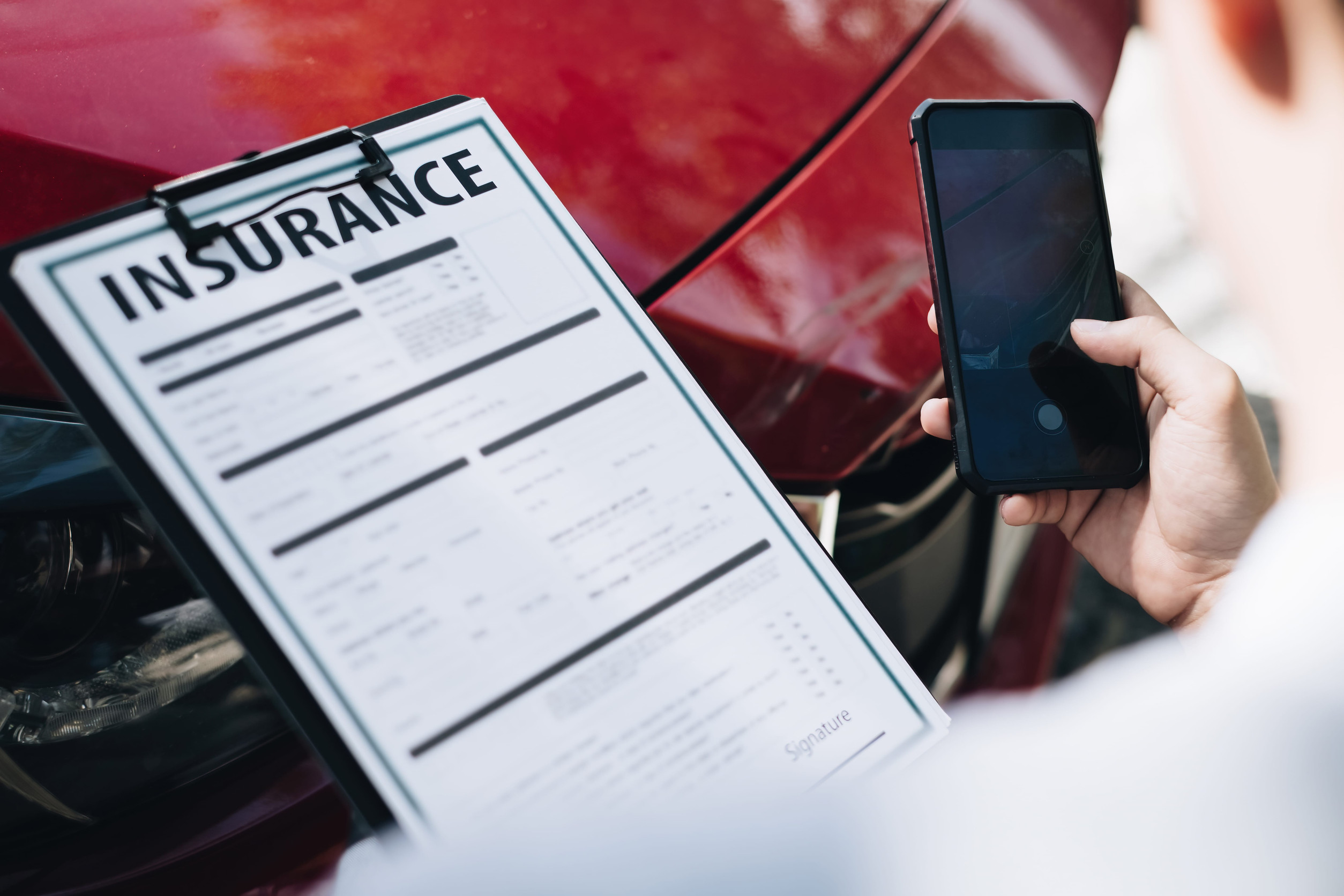 A person holds a smartphone to take a photo of a damaged red car while holding an auto insurance claim form on a clipboard, documenting the accident for insurance coverage.