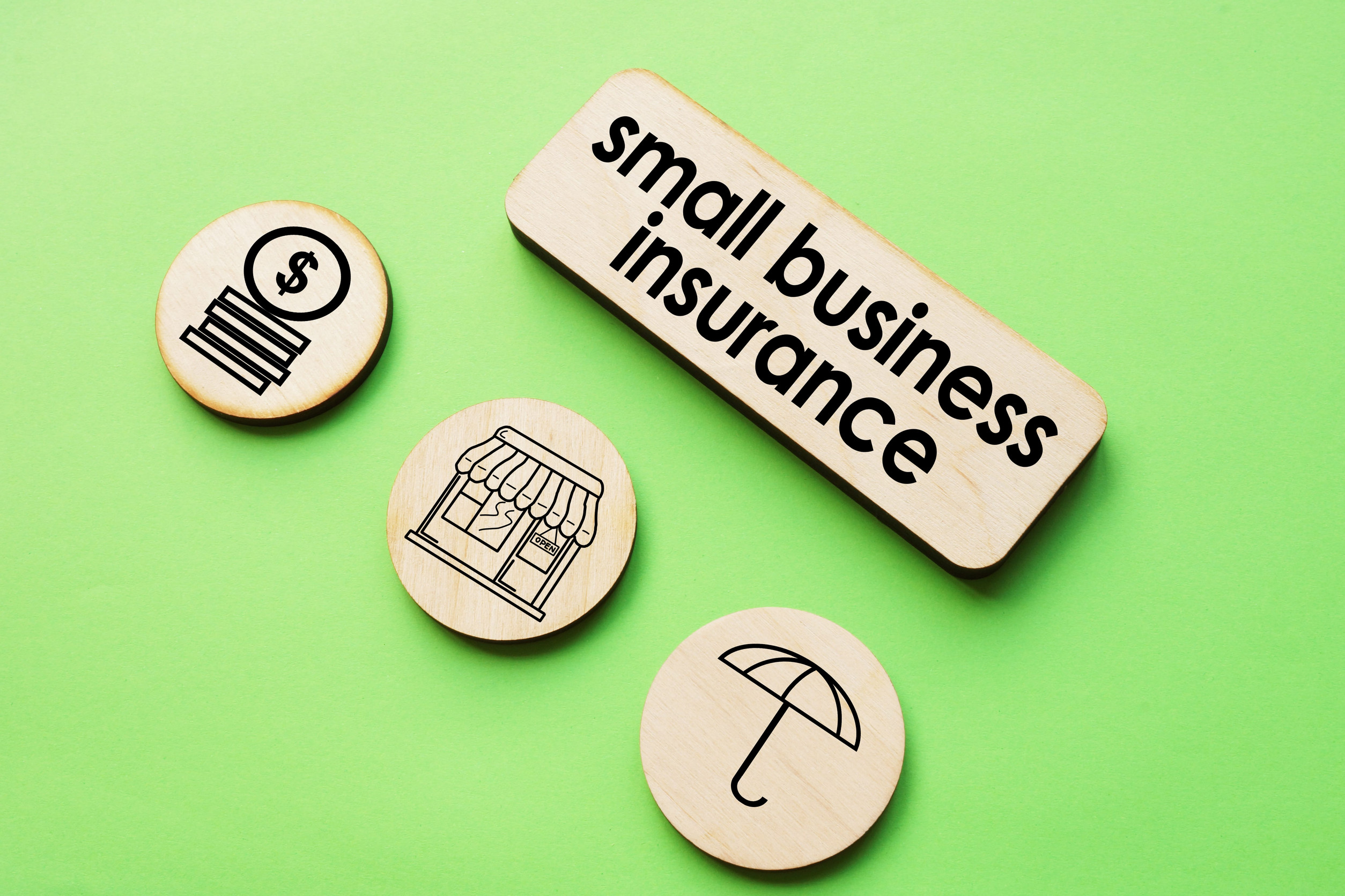 Wooden tokens on a green background show icons of money, a shop, and an umbrella, with a larger sign reading “small business insurance,” reflecting the importance of Business Insurance for both traditional storefronts and Home-Based Business owners.