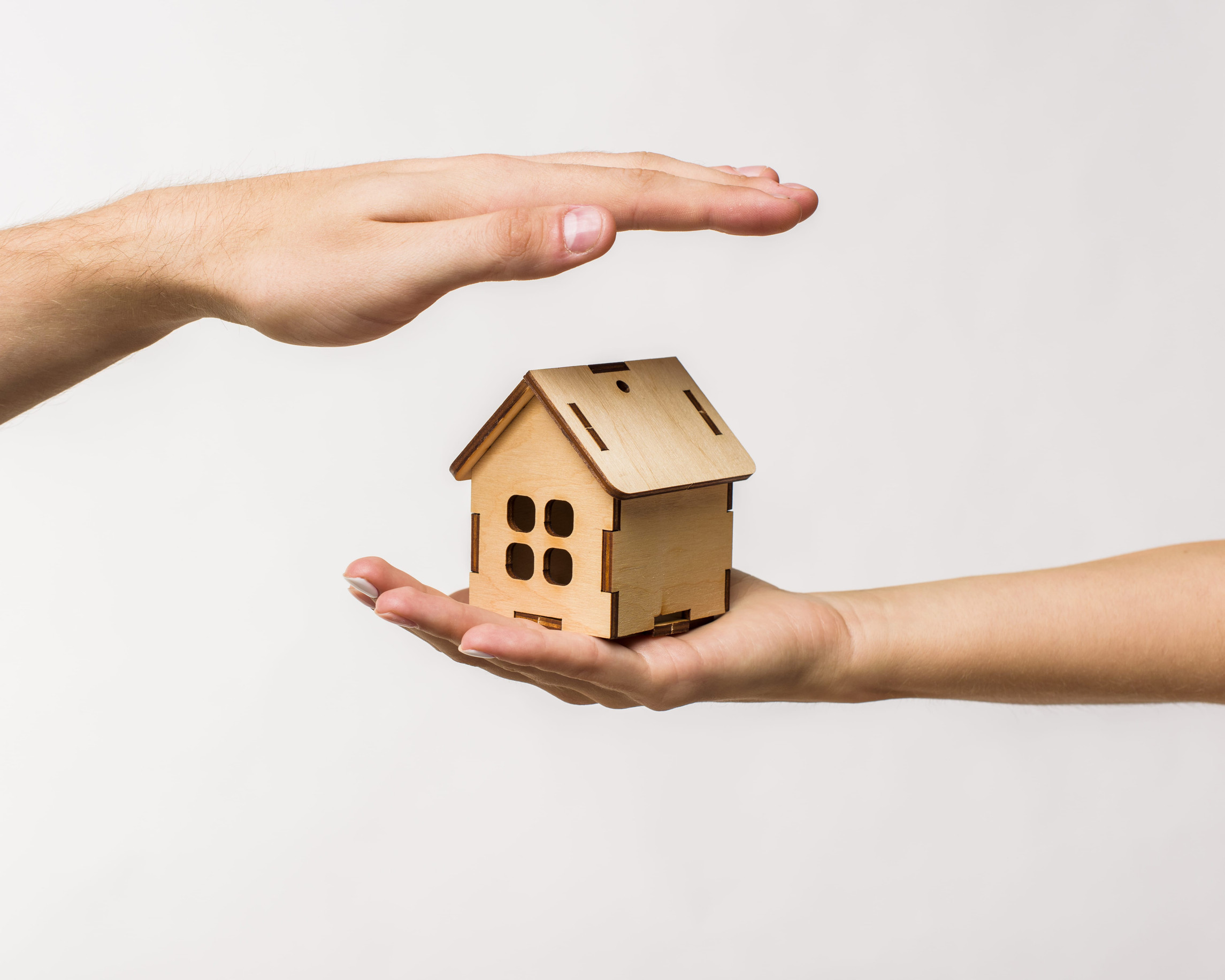 Two hands surround and protect a small wooden house model, one hand above and one below, against a plain white background—illustrating the peace of mind that comes with reliable Minnesota Cabin Insurance as you prepare to close for winter.