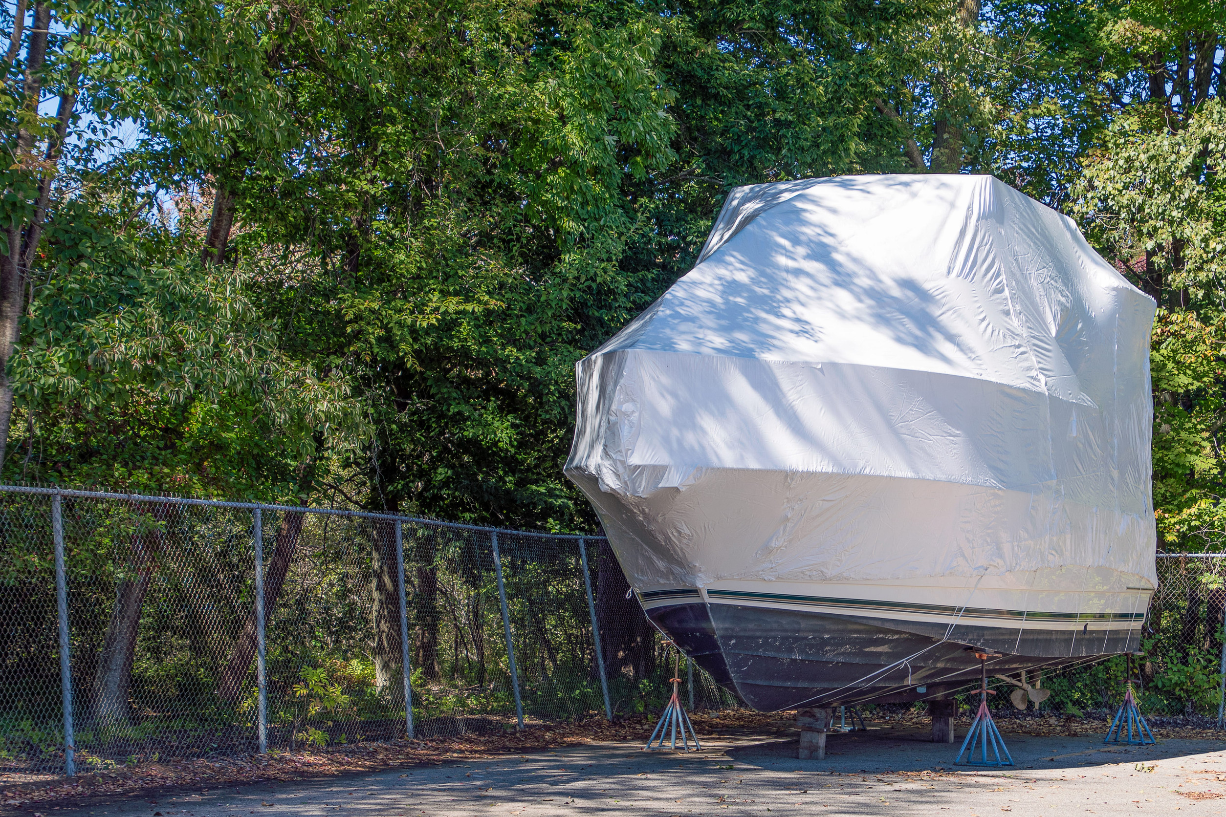 A boat covered in white plastic is stored on supports in a paved lot next to a chain-link fence, with green trees in the background—an example of proper winter storage that helps protect your investment and may even be required for certain boat insurance policies.