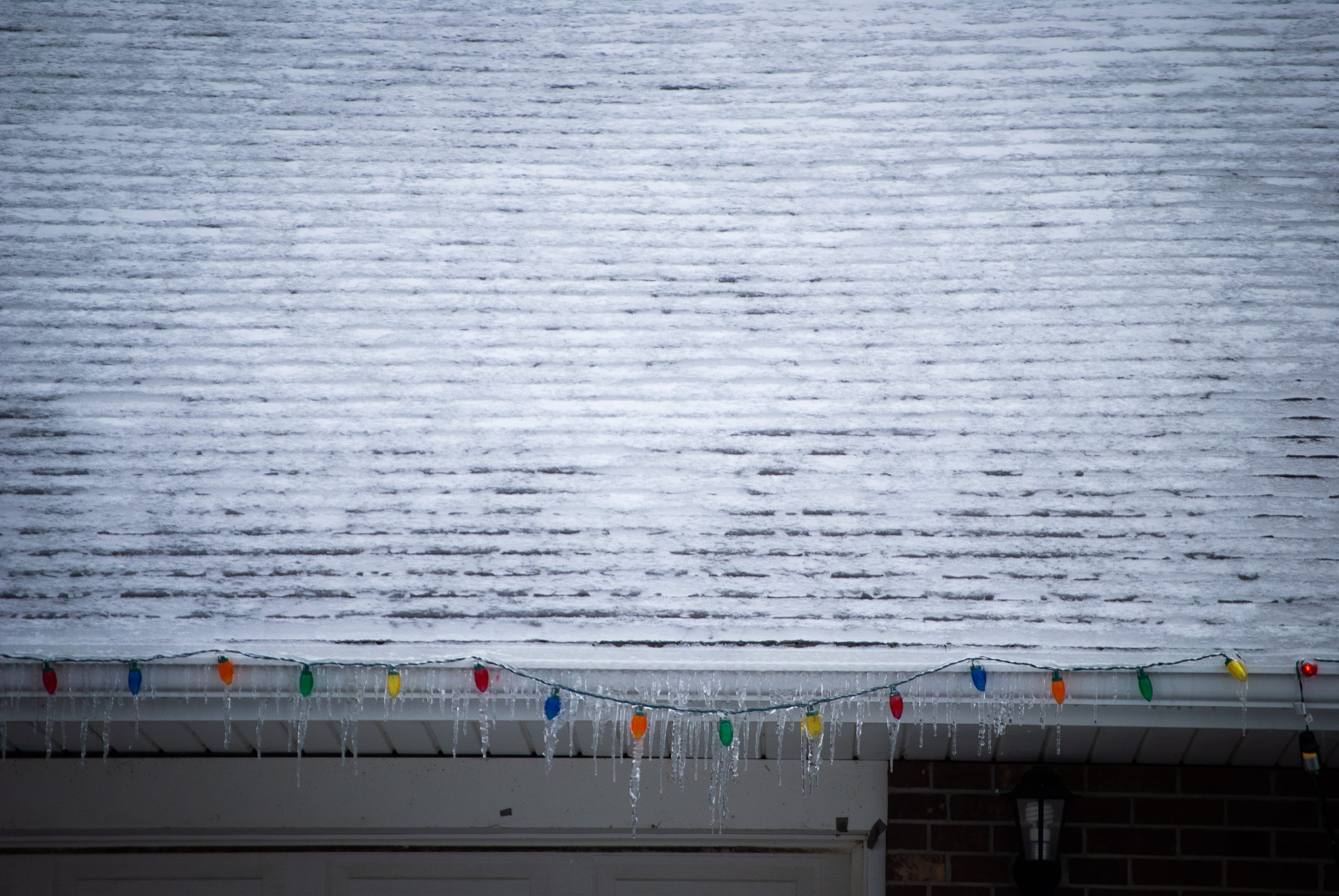 A snow-covered roof edge lined with colorful Christmas lights and hanging icicles that may require roof ice dam coverage.