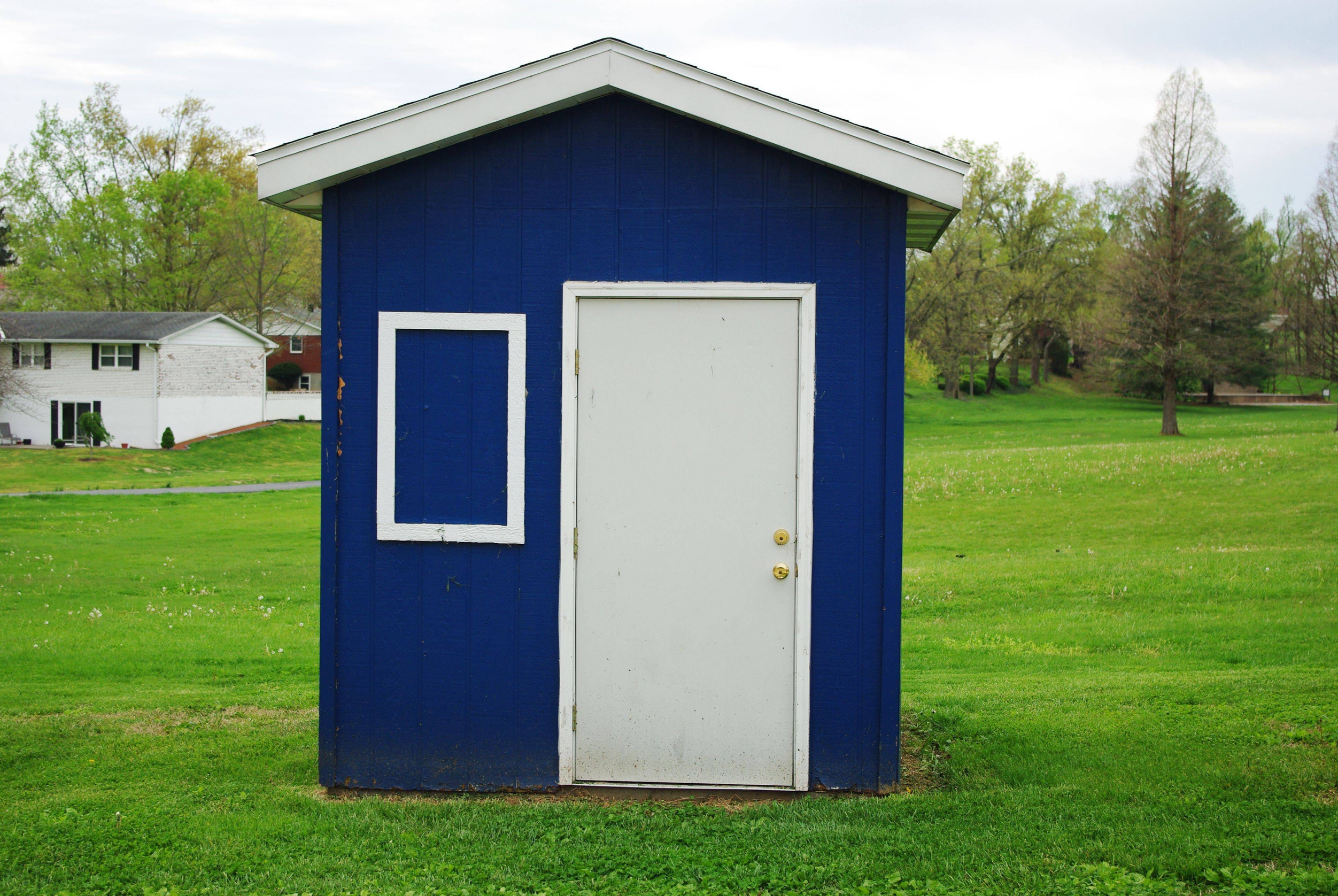 Small blue shed with a white door and an empty white window frame in a grassy field that might be protected by detached garage insurance.
